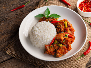 Rice topped with stir fried crispy pork with red curry in white plate on wooden table background. Thai Food