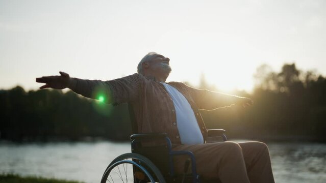 Senior man in wheelchair enjoys freedom. Paralysis male with special need spreading arms on sides happy breathing fresh air walking in city park. Living with physical disability in old age concept.