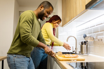 A dark-skinned couple is doing housework in the kitchen of their apartment. The African man is...