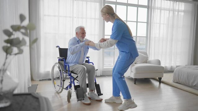Nurse talking senior man in wheelchair. Female welfare physical therapist looks after senior male, listening, providing medical care at clinic. Living with legs paralysis, special needs people.