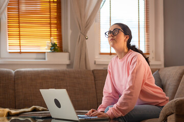 Pensive young Caucasian woman wearing glasses is sitting on couch and using her laptop. Cozy...