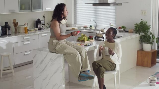 Medium long shot of young diverse women talking about university and eating snacks while resting at weekend morning