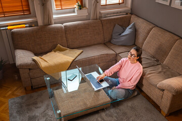 Top view of young boring Caucasian woman wearing glasses is sitting near sofa and using laptop....