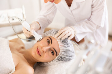 Young woman undergoing device-driven cosmetic facial treatment using high frequency ultrasound attachment stimulating opening of skin pores for deep penetration of moisturizers and nutrients