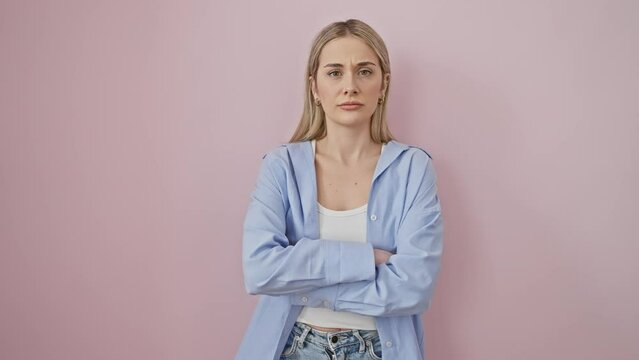 Disapproving blonde, a young woman skeptically crossing arms over pink backdrop, wearing emotion of nervousness and negativity on her face.