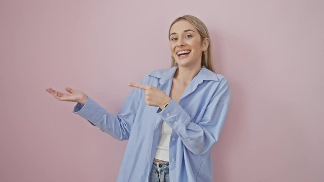 Stunned and beautiful blonde woman pointing, presenting with hand while wearing shirt, ecstatically smiling against vibrant pink wall, exuding confidence.