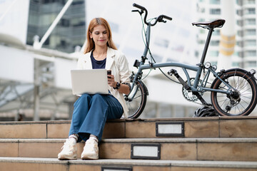 Confident businesswoman ride bicycle sit outside in downtown working using laptop. Entrepreneur...