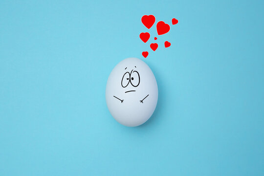Funny white egg with face feeling on blue background with red hearts above it. Copy space. Emoticons concept. Art collage