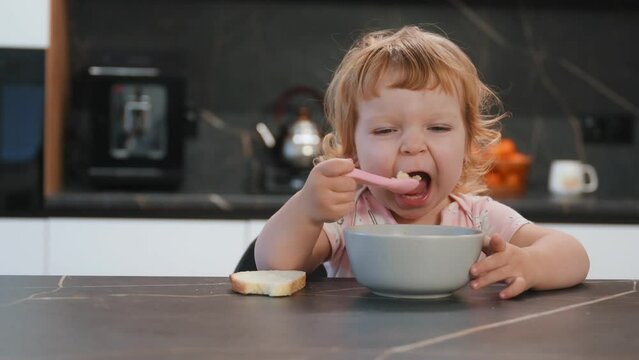 A small beautiful girl with big blue eyes is eating soup with a spoon in a home by herself. Development of children. Healthy nutrition in childhood.