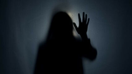A dark silhouette of a woman behind a mat curtain, illuminated by a beam of light. The woman...