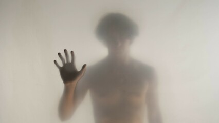 Blurred silhouette of a man with a naked torso behind a frosted curtain or glass. The man touches the transparent curtain with two palms and looks into the camera.