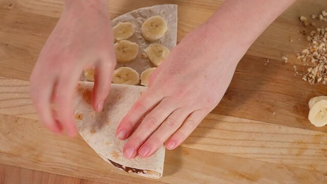 Girl preparing healthy vegan breakfast with chocolate spread pita bread with nuts and bananas, wooden table. The round tortilla is divided into four fillings; the girl&rsquo;s hands fold the tortilla into a