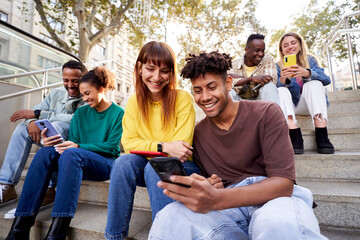 Multiracial young friends sitting on stairs hanging out, using phone to send messages while being...