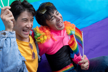 Two gay men sitting on rainbow flags on the ground, celebrate by waving flags to welcome the pride...