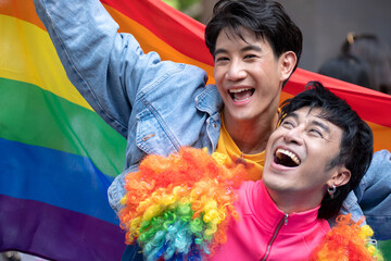 Two gay men sitting on rainbow flags on the ground, celebrate by waving flags to welcome the pride...