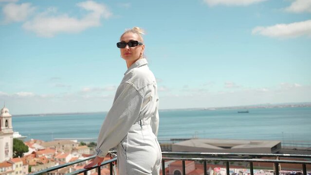 Rear view of excited young female tourist on viewpoint Portas do Sol in Lisbon. Beautiful young woman in sunglasses looking at camera while enjoying the view of sea and town. Lady on summer vacation.