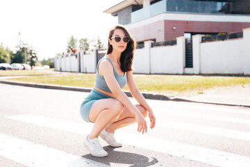 Young beautiful confident female in trendy summer blue cycling shorts and tank top clothes. Carefree woman posing in street. Positive model having fun. Cheerful and happy. Sits on asphalt
