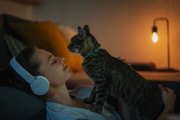 Young smiling happy woman in headphones sitting with a gray cat pet in her arms at home in the night
