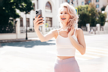 Young beautiful smiling blond hipster woman in trendy summer clothes. Carefree female posing in the street at sunny day. Positive model outdoors at sunset. Cheerful and happy. Takes selfie photos