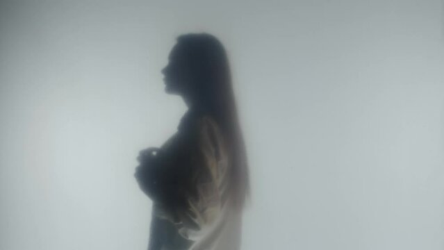 Side view of silhouette of blonde woman unbuttoning buttons on white blouse. Profile portrait of a woman in steam or fog behind a clear frosted glass or curtain close up.