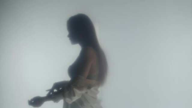 Side view of silhouette of a blonde woman removing her white blouse. Profile portrait of a seminude woman in steam or fog behind clear frosted glass or curtain close up.