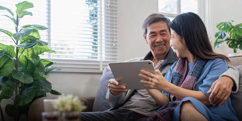 Happy elderly asian couple using tablet sit on sofa doing ecommerce shopping online on website and...