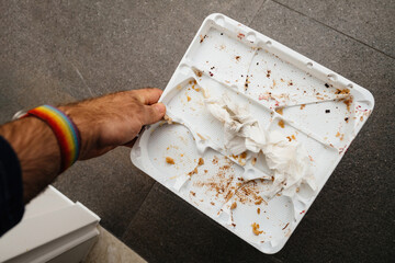 A male hand holds an empty plastic cake tray above a stone background, showcasing small traces and crumbs, remnants of an afterparty