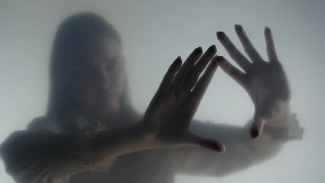 Silhouette of ghost woman in fog behind frosted curtain or glass. Woman's hands touching the glass in close up. Concept of afterlife, otherworld, ghosts.