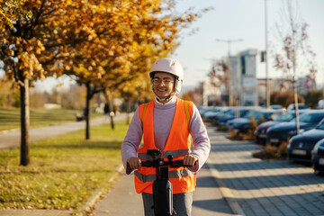 Obraz premium A careful young woman in vest and helmet is driving safely an electric scooter on a street.