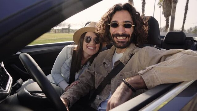 Young Latin attractive couple in love pose smiling for photo inside convertible car on sunny day. Cheerful millennial people with sunglasses enjoying summer vacation together. Free time outdoors 