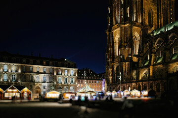 An evening view of a vibrant outdoor market beside a Gothic cathedral in Strasbourg, brilliantly illuminated under the starry night sky