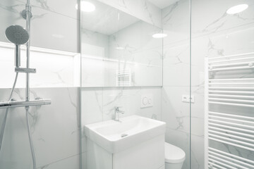 A Modern, bright bathroom with white marble walls, a shower, sink, toilet, and a heated towel rack....