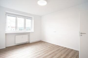 A Minimalist, bright room with white walls, a wooden floor, three windows, and an opened door. It’s clean and empty.