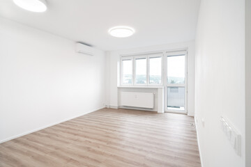 A Bright, empty room with white walls, a large window, wooden floor, and a modern ceiling light. An air conditioner is mounted on the wall.