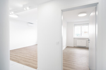 A Bright, modern apartment with white walls and wooden flooring. A window allows natural light to illuminate the minimalist space. A doorway leads to another room. There is AC interior unit on wall.