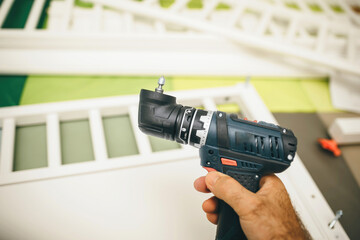 A persons hand firmly grips a cordless electric drill with a bit attached in a construction site environment, indicating an ongoing building or repair task - assembling furniture