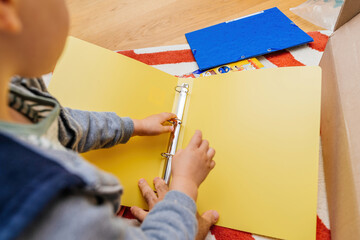 A young toddler is securing papers within a yellow binder on the floor of a vibrant classroom, surrounded by stationary and learning materials.