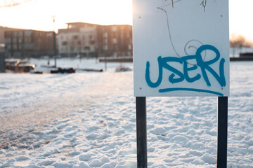 User graffiti sprayed onto a white sign in a park. Winter morning atmosphere with snow-covered park...