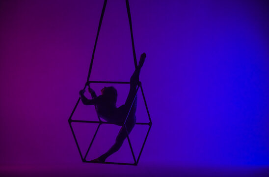 Silhouette of female aerial gymnast isolated on blue purple neon background. Female acrobat performing in the air on a cube, showing her flexibility.
