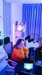 Young Caucasian woman professional gamer wearing orange hoodie sits on a chair with a hand under her chin, feeling upset, sad when losing esports competition. Concept of pain from gaming and esports.