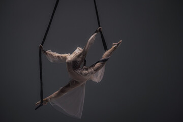 Girl aerial gymnast demonstrates stretching in twine on acrobatic trapeze. Acrobatic athlete performs hanging from a height in a dark studio.