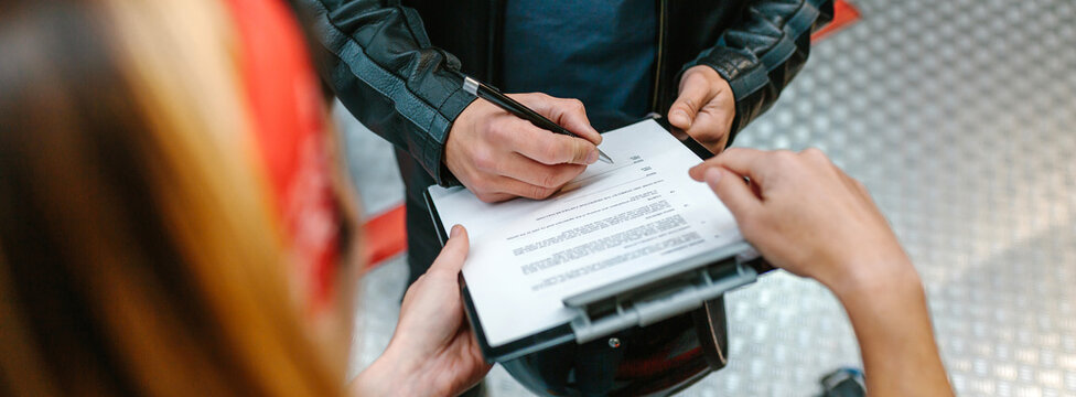 Close up of unrecognizable female mechanic with red hair bandana holding a clipboard while biker man wearing leather jacket signing insurance policy