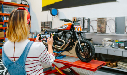 Unrecognizable female mechanic taking photo with cellphone to custom motorcycle repaired over platform on garage. Selective focus on phone screen. - Powered by Adobe