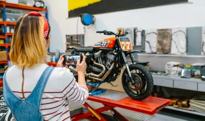 Papier Peint photo autocollant Moto Unrecognizable female mechanic taking photo with cellphone to custom motorcycle repaired over platform on garage. Selective focus on phone screen.