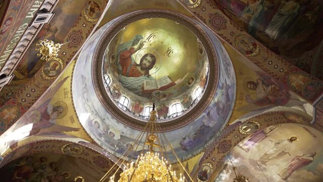 The majestic dome of an Orthodox church, adorned with the Christ Pantocrator fresco, surrounded by ornate murals and a hanging chandelier.
