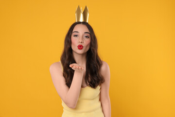 Beautiful young woman with princess crown blowing kiss on yellow background