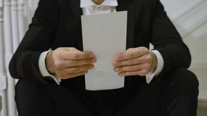 man in a black suit holding a white envelope. He is seated and focused on the content, possibly preparing to read a letter or an invitation. conveys a sense of anticipation and formality