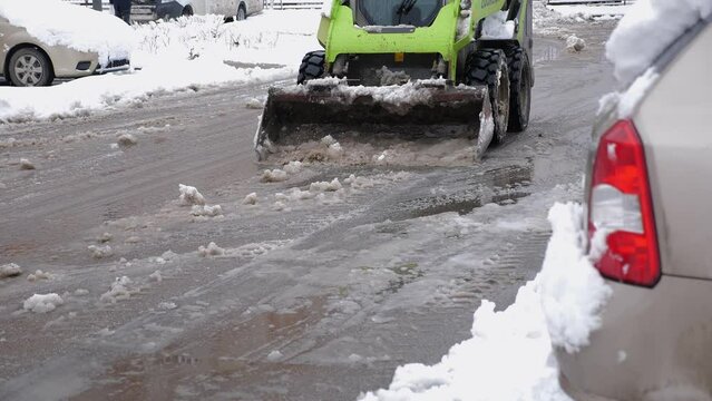 A close-up of a small tractor with a bucket cleans the road in the parking lot from slush and snow. Municipal transport clears roads from snow in the courtyard of high-rise buildings. 