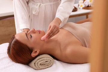 Fototapeta na wymiar Young woman receiving facial massage with rose quartz gua sha tool in beauty salon, closeup