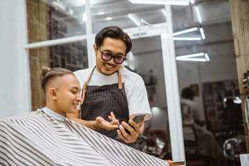 asian barber holding phone and showing it to his customer asking for feedback with friendly expression